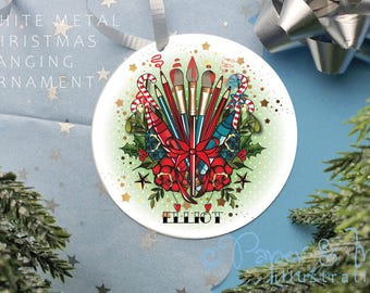 May include: White metal Christmas ornament with a green background and a red bow. The ornament features a collection of paintbrushes, pencils, and candy canes. The name "Elliot" is written in black lettering at the bottom of the ornament.