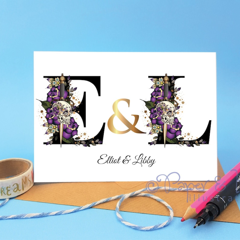 May include: A white card with a black and gold design featuring the letters "E" and "L" with a skull and dagger motif. The text "Elliot & Libby" is printed below the letters.