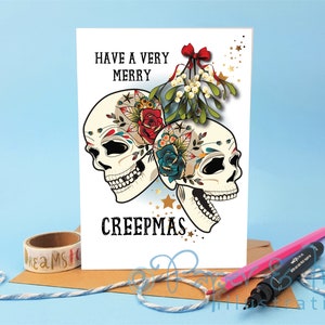 May include: A white card with a festive message that reads "Have a Very Merry Creepmas". The card features two colorful sugar skulls with floral designs and a sprig of mistletoe with a red bow.