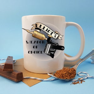 May include: White ceramic mug with a black and gold tattoo machine design. The mug has the text "Elliot's Weapon of Choice" printed on it.