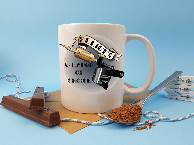 May include: White ceramic mug with a black and gold tattoo machine design. The mug has the text "Elliot's Weapon of Choice" printed on it.