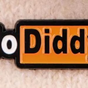 May include: A rectangular black and orange pin with the words "No Diddy" in white and black lettering. The pin is set against a textured, off-white background.