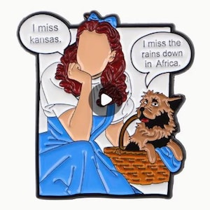 May include: Enamel pin featuring a cartoon illustration of a character in a blue dress with a dog in a basket. The character has red hair and a blue bow. Text bubbles read, "I miss kansas" and "I miss the rains down in Africa."
