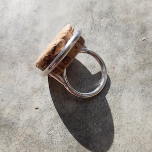 May include: A silver ring with a natural cork feature. The ring has a simple, modern design with a circular band and a cork top. The cork is light brown, and the metal is shiny silver.