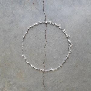 Könnte beinhalten: Eine silberne Halskette mit einer zarten Kette und einem einzigartigen, strukturierten Design. Die Halskette liegt auf einer grauen Betonfläche.