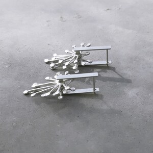 May include: A pair of silver earrings with a delicate, starburst design. The earrings are made of a thin, silver bar with a cluster of small, silver circles radiating out from the center.