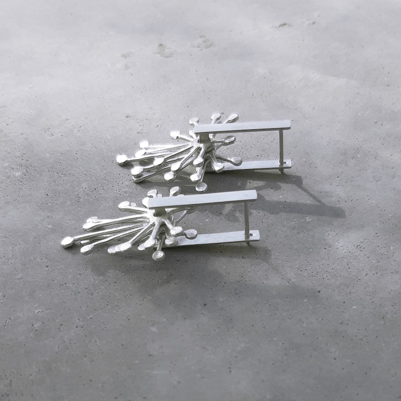 May include: A pair of silver earrings with a delicate, starburst design. The earrings are made of a thin, silver bar with a cluster of small, silver circles radiating out from the center.