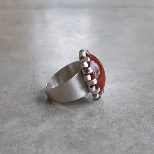 May include: A silver ring with a red gemstone set in a silver bezel. The bezel is decorated with a textured design.