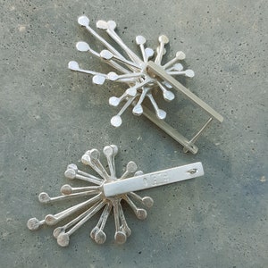 May include: Two silver earrings with a starburst design. The earrings have a silver bar with a stamp that reads "925" on the back.