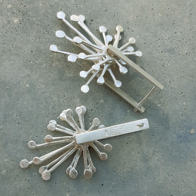May include: Two silver earrings with a starburst design. The earrings have a silver bar with a stamp that reads "925" on the back.
