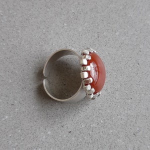 May include: A silver ring with a red carnelian stone set in a decorative silver bezel. The ring is on a gray surface.
