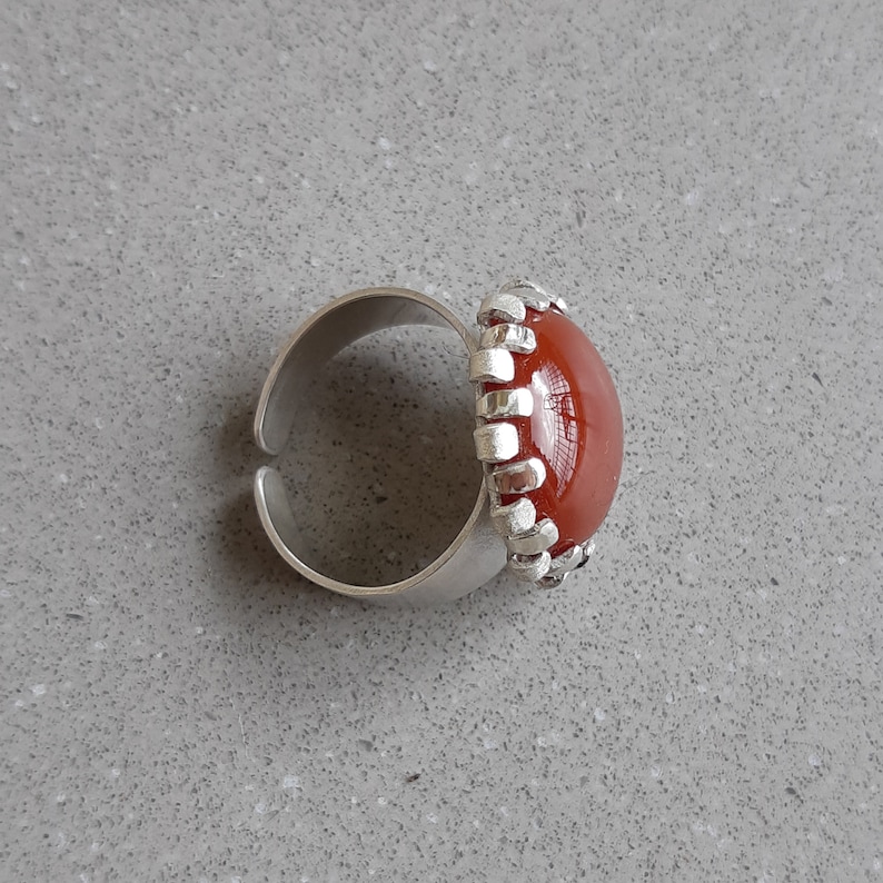 May include: A silver ring with a red carnelian stone set in a decorative silver bezel. The ring is on a gray surface.