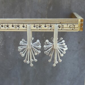 May include: A pair of silver earrings with a delicate, starburst design. Each earring features a thin, vertical bar with a cluster of small, rounded shapes radiating outwards.