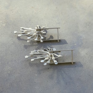 May include: A pair of silver earrings with a delicate, geometric design. Each earring features a cluster of small, rounded shapes radiating outwards from a central point. The earrings are attached to a simple, straight post.