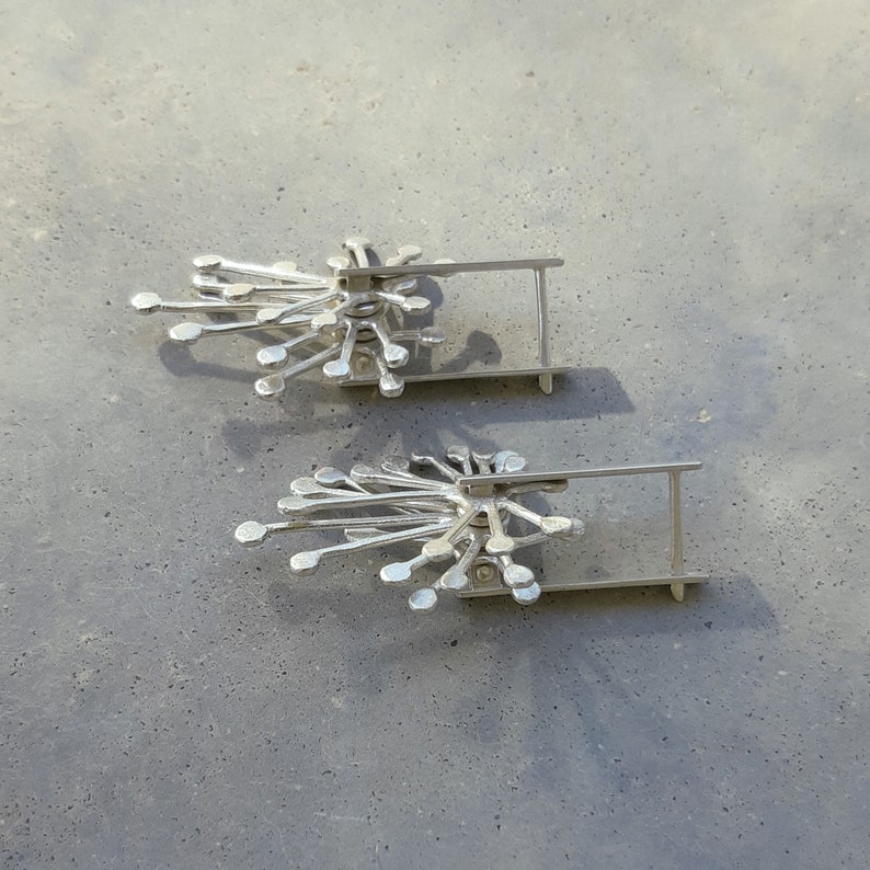 May include: A pair of silver earrings with a delicate, geometric design. Each earring features a cluster of small, rounded shapes radiating outwards from a central point. The earrings are attached to a simple, straight post.