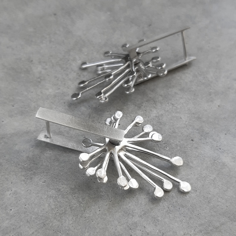 May include: A pair of silver earrings with a geometric design. Each earring features a starburst shape with multiple points radiating outwards. The earrings are attached to a silver bar.