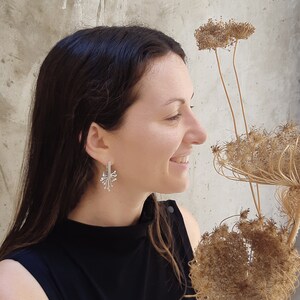 May include: A woman with long dark hair is wearing silver earrings with a delicate, intricate design. The earrings are shaped like a burst of light or a flower.