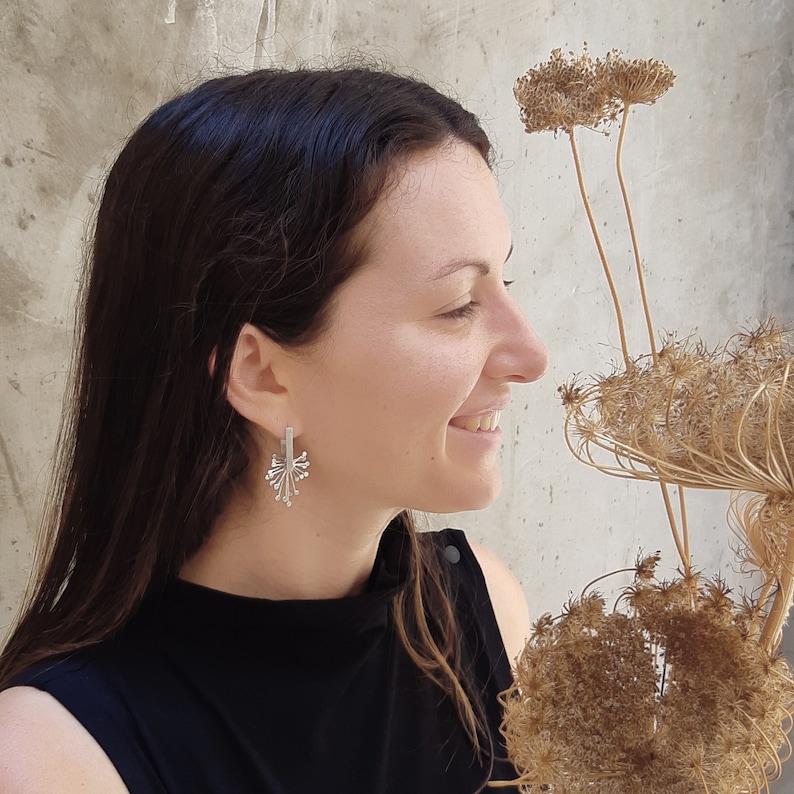 May include: A woman with long dark hair is wearing silver earrings with a delicate, intricate design. The earrings are shaped like a burst of light or a flower.