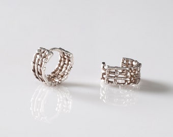 May include: A pair of silver hoop earrings with a geometric, woven design. The earrings are made of a thick, textured metal.