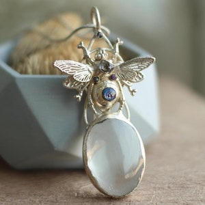 May include: Silver pendant featuring a detailed insect design with iridescent blue and purple gemstones. The pendant is attached to a silver chain and hangs from a white oval-shaped stone.