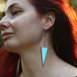 May include: Long, geometric earrings with a light blue, triangular pendant on a silver chain. The earrings are worn by a person with red hair and a black top. The background is blurred.