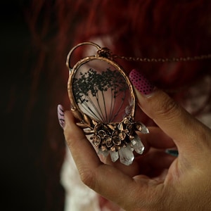 May include: An oval pendant necklace featuring a clear resin centre with a black pressed botanical silhouette. The frame is made of copper-toned metal with decorative floral accents at the base, complemented by small, clear, pointed crystal shards.