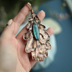 May include: Copper pendant necklace shaped like an oak leaf, with a central oval blue-green gemstone. The leaf has detailed veining and a textured surface, suspended from a delicate copper chain. A unique piece of jewellery.