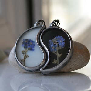 May include: A yin yang pendant necklace with a white and black background. Each half of the pendant features a pressed blue flower.