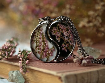May include: A silver-toned pendant necklace with a yin-yang design. One side is white with pink flowers, the other black with pink flowers. The necklace rests on a vintage book with a pink cover, with sprigs of pink flowers and a clear crystal nearby.