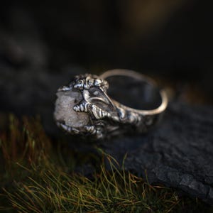 May include: A silver ring featuring a raw, light-colored stone held in a claw-like setting. The ring has intricate, detailed metalwork and a polished finish. The ring is set against a dark, natural background.