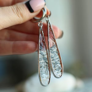 Op de afbeelding: Een paar lange, druppelvormige oorbellen. De oorbellen hebben een zilverkleurig metalen frame en zijn gevuld met een getextureerd, helder materiaal dat licht reflecteert. De binnenkant heeft een roodbruine tint. De oorbellen hangen aan zilveren creolen.