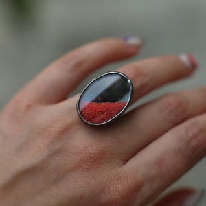 以下が含まれることがあります： 透明なドーム型のトップが付いた、楕円形のシルバーリング。リングの内側には、暗い部分と小さな赤いビーズのクラスターがあります。リングは指に装着されており、そのユニークなデザインと色のコントラストが際立っています。
