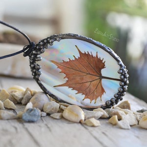 May include: An oval pendant featuring a dried brown maple leaf encased in iridescent resin, framed by a textured dark metallic border. The pendant hangs from a thin black cord and includes the text ZebraArtStudio etched on the surface.