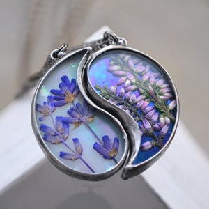 May include: A silver yin yang necklace with two halves, each containing pressed flowers. One half features purple flowers on a white background, the other half features pink flowers on a blue background.
