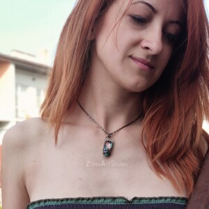 May include: A silver chain necklace with a pendant featuring a small, rectangular, green and pink glass piece with a floral design. The text "ZEBRA ART STUDIO" is visible below the pendant.