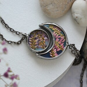 May include: A silver chain necklace with a crescent moon pendant featuring pressed flowers in shades of pink and purple. The pendant is set against a blue and green background.