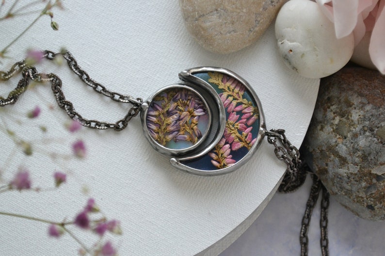 May include: A silver chain necklace with a crescent moon pendant featuring pressed flowers in shades of pink and purple. The pendant is set against a blue and green background.