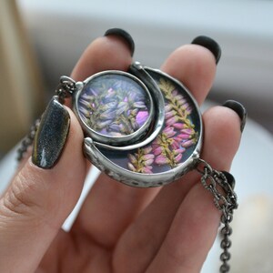 May include: A silver crescent moon pendant necklace with a round glass charm featuring pressed purple and pink flowers. The necklace has a silver chain.