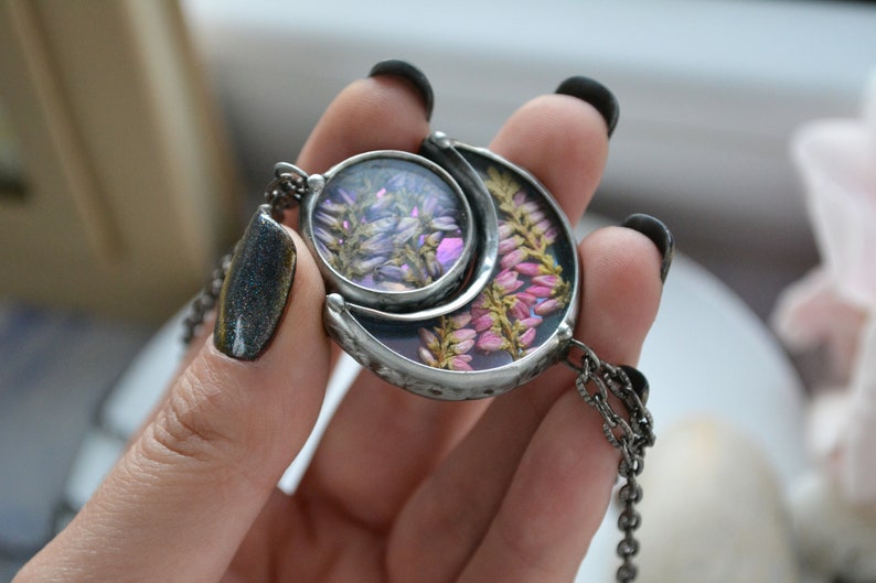 May include: A silver crescent moon pendant necklace with a round glass charm featuring pressed purple and pink flowers. The necklace has a silver chain.