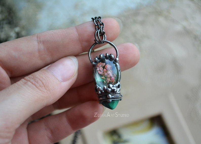 May include: A silver chain necklace with a pendant featuring a green malachite stone and a clear glass dome containing dried pink flowers. The pendant is surrounded by silver wire and beads. The text "ZEBRA ART STUDIO" is visible at the bottom of the image.