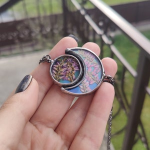May include: A silver crescent moon necklace with a round pendant featuring pressed flowers in shades of pink and purple. The pendant is set in clear resin and has a blue iridescent background.