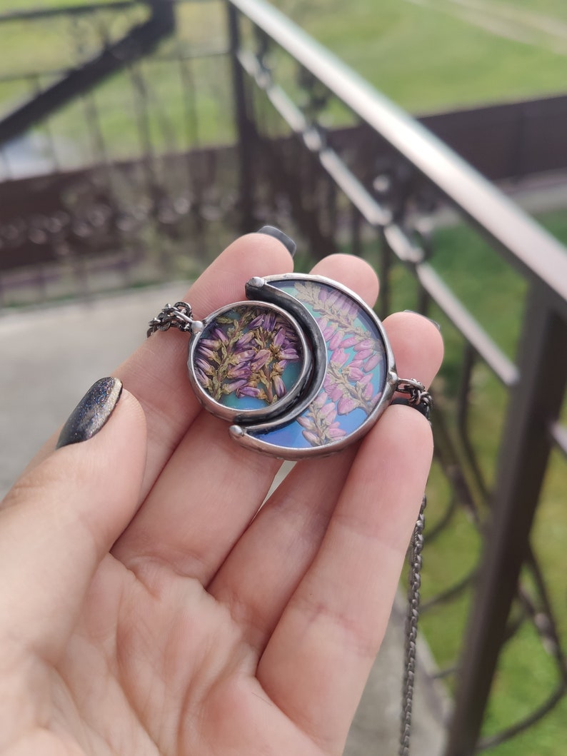 May include: A silver crescent moon necklace with a round pendant featuring pressed flowers in shades of pink and purple. The pendant is set in clear resin and has a blue iridescent background.