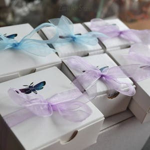 May include: A set of eight white gift boxes with purple and blue sheer ribbon bows. Each box has a different design featuring a butterfly or a bee. The boxes are arranged in two rows of four.