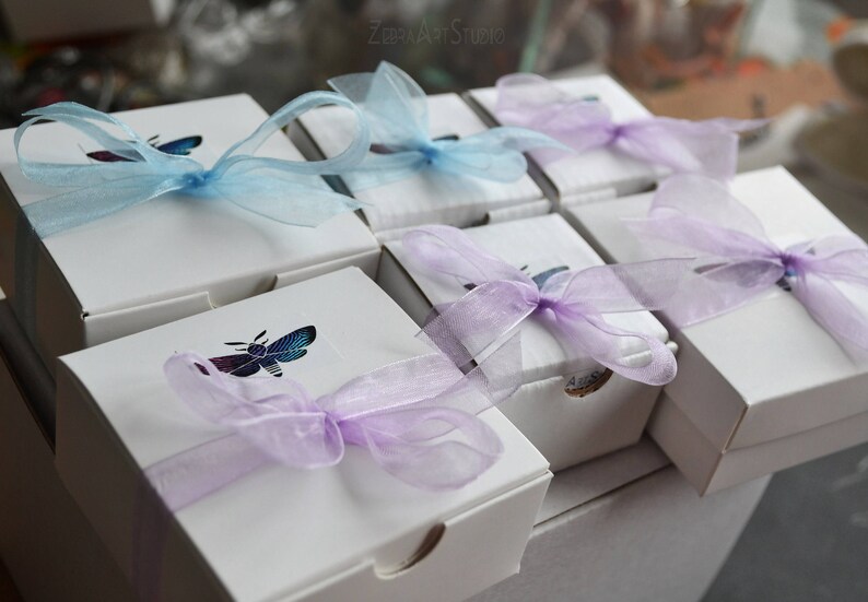 May include: A set of eight white gift boxes with purple and blue sheer ribbon bows. Each box has a different design featuring a butterfly or a bee. The boxes are arranged in two rows of four.