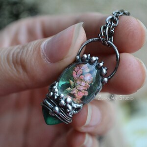 May include: A silver chain necklace with a pendant featuring a green and pink flower encased in resin. The pendant is surrounded by silver beads.