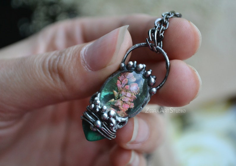 May include: A silver chain necklace with a pendant featuring a green and pink flower encased in resin. The pendant is surrounded by silver beads.