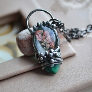 May include: A silver necklace with a green and silver pendant featuring a pressed flower encased in resin. The pendant is shaped like a teardrop and has a delicate, intricate design.