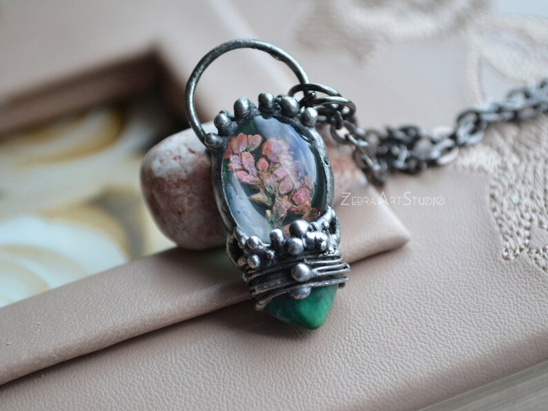 May include: A silver necklace with a green and silver pendant featuring a pressed flower encased in resin. The pendant is shaped like a teardrop and has a delicate, intricate design.