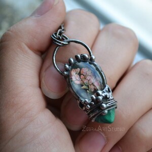 May include: A silver pendant necklace with a green malachite stone and a clear resin oval containing pressed pink flowers. The pendant is attached to a silver chain.