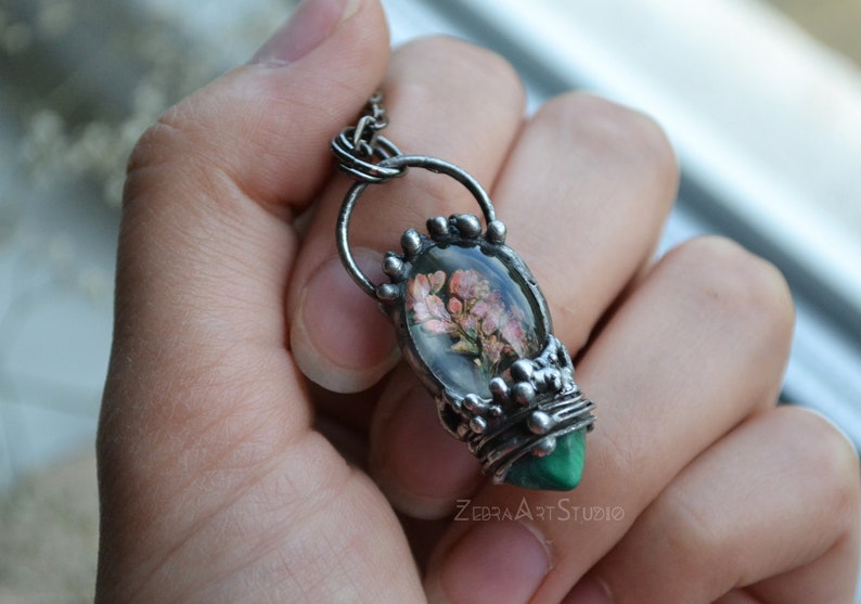 May include: A silver pendant necklace with a green malachite stone and a clear resin oval containing pressed pink flowers. The pendant is attached to a silver chain.